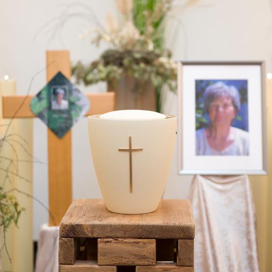 Urne mit Kreuzsymbol auf Holzständer, umgeben von Pflanzen, Kerzen und einem Foto in einem festlichen Raum.