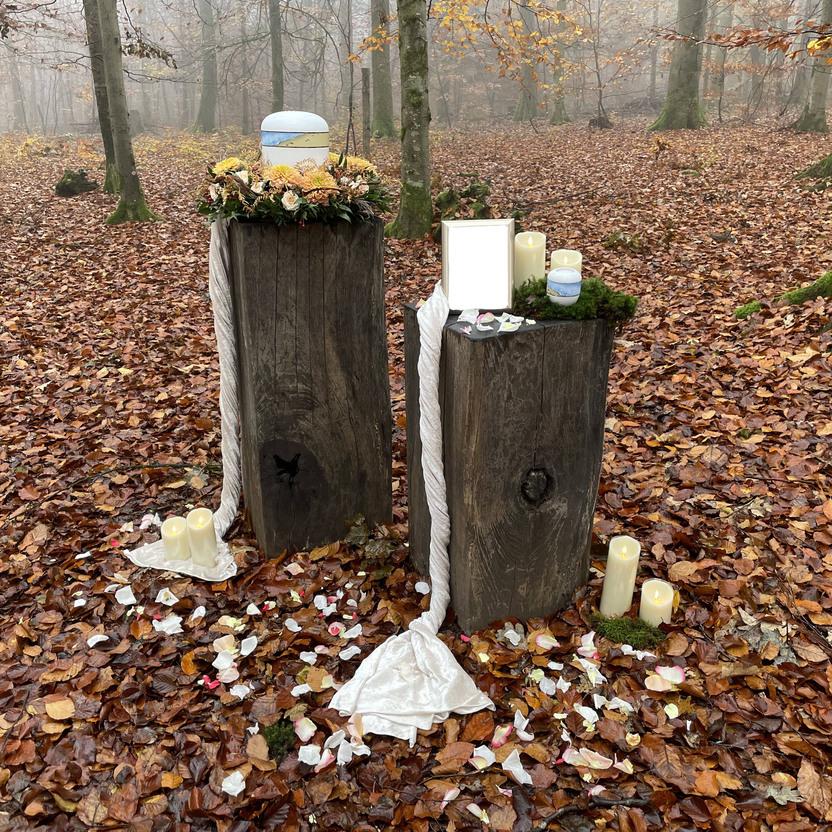 Zwei Holzskulpturen stehen im nebligen Wald, geschmückt mit Blumen, Kerzen und Blütenblättern auf dem Laub.