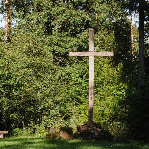 Holzkreuz auf einem Steinhaufen in einer Waldlichtung, umgeben von hohen Bäumen und einem Holzbank.