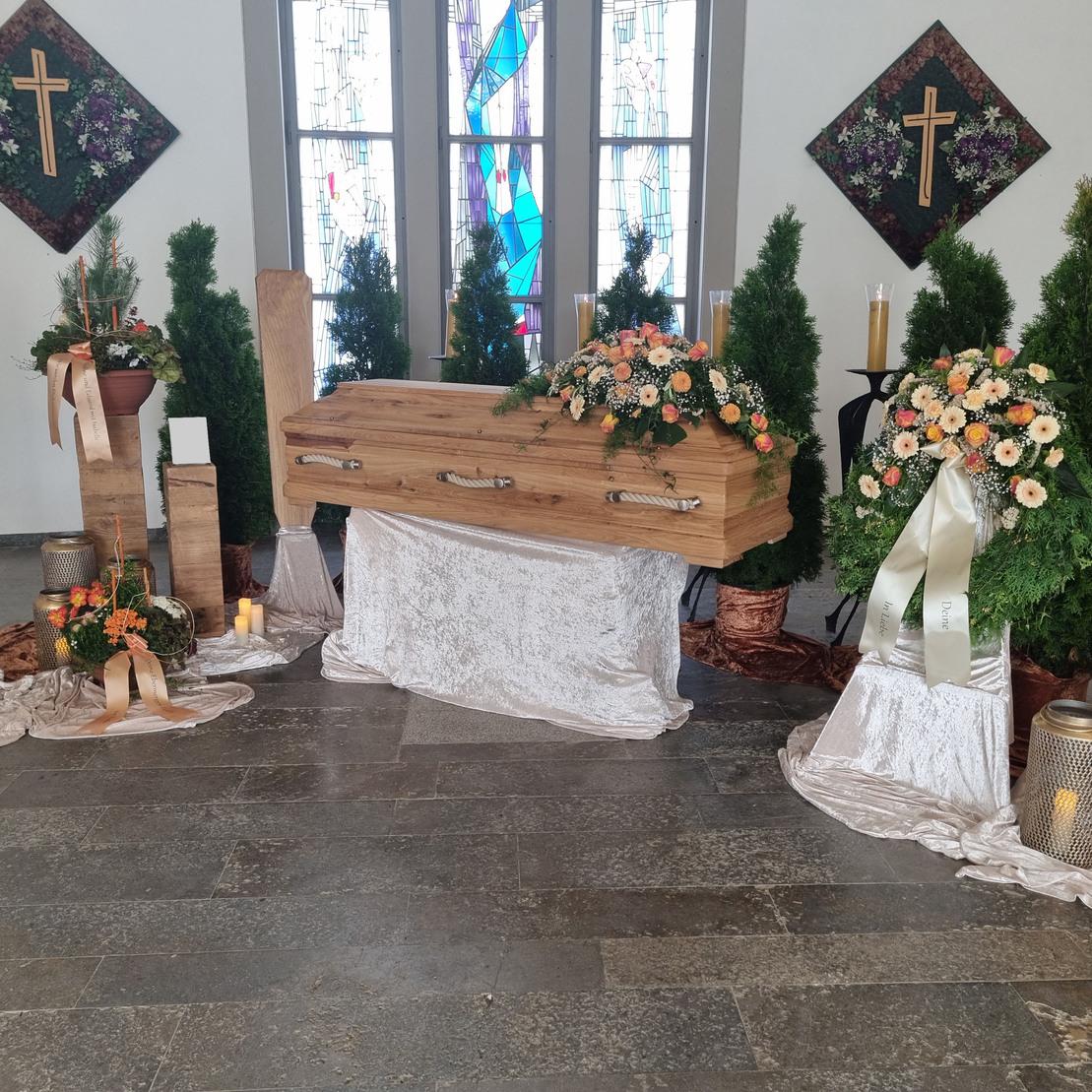 Dekorierte Trauerstätte mit einem Holzsarg, Blumenarrangements und Kerzen auf einem eleganten Tuch.