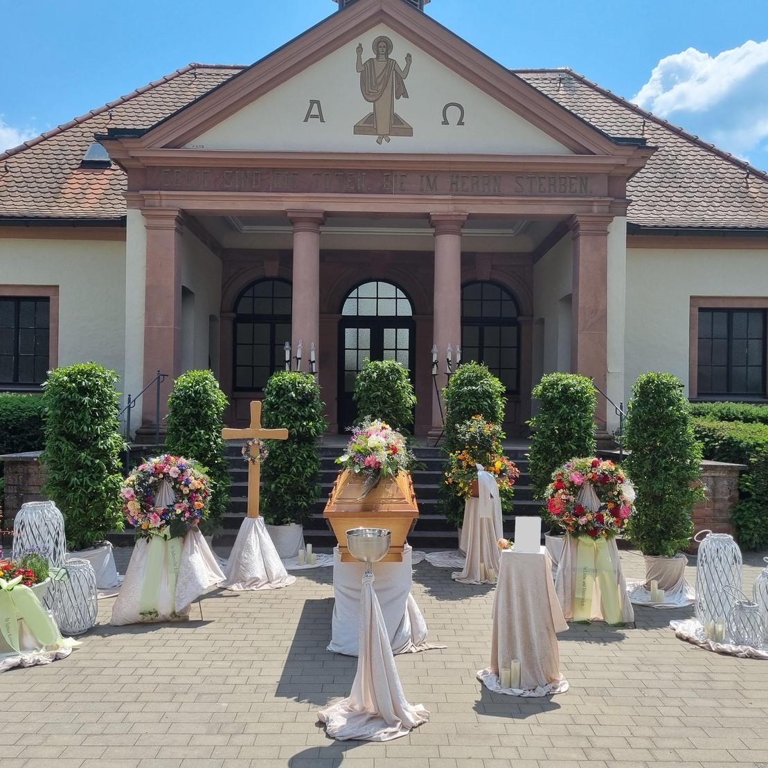 Feierliche Trauerzeremonie im Freien mit einem geschmückten Sarg, Blumenkränzen und einer Kapelle im Hintergrund.