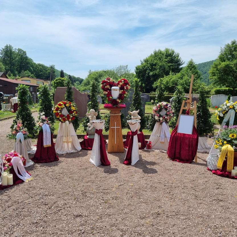 Dekorierte Grabstelle mit Blumenarrangements, Kerzen und einem Holzständer auf einem Friedhof bei sonnigem Wetter.
