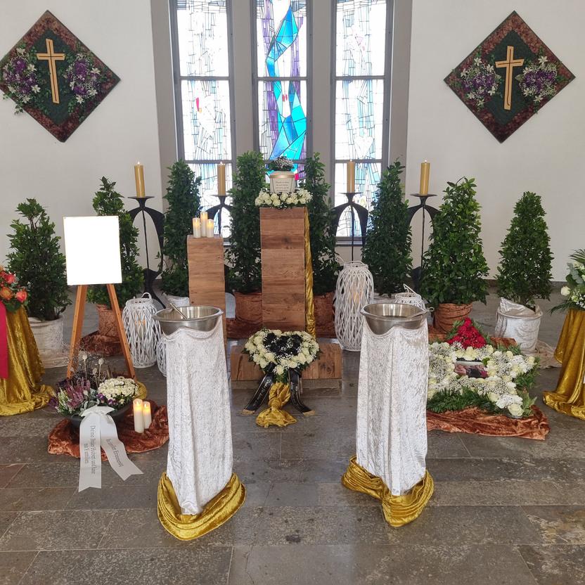 Dekorierter Raum mit Blumen, Kerzen und einem Kreuz, umgeben von Bäumen und einem Holzpodest für eine Trauerfeier.