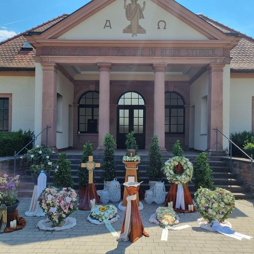 Blumenarrangements und Kränze vor einer Kirche mit Säulen und einem Walmdach unter blauem Himmel.