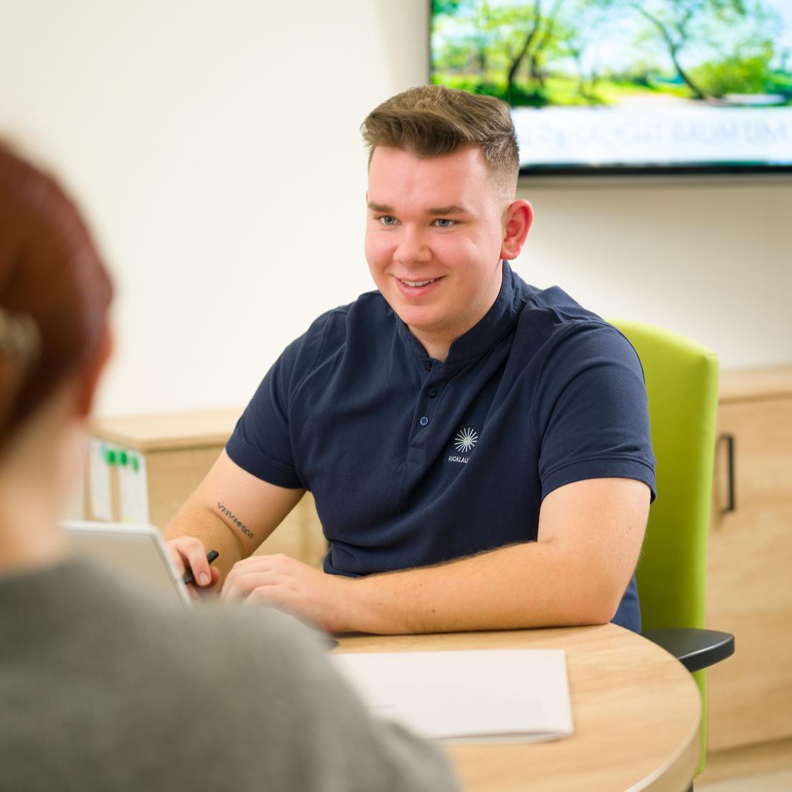 Junger Mann in blauen Poloshirt sitzt an einem Tisch und spricht mit einer Person, die zu ihm blickt. Hintergrund mit Bildschirm.
