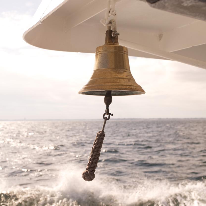 Goldene Schiffsglocke, aufgehängt an einem Boot, mit schäumenden Wellen im Hintergrund und einem bewölkten Himmel.