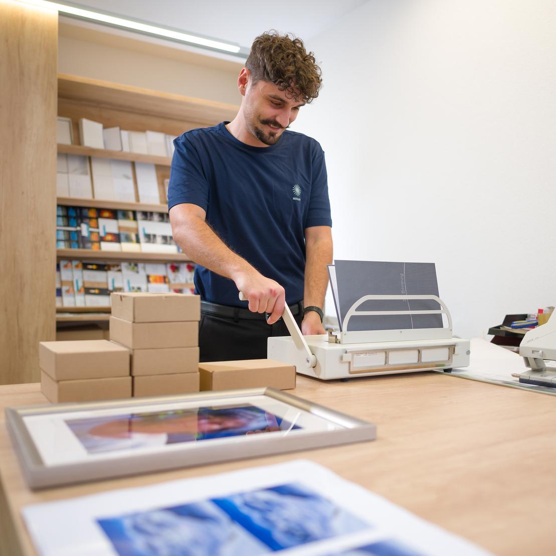 Mann schneidet Papiere an einem Tisch in einem Geschäft, im Vordergrund sind gestapelte Kartons und Bilderrahmen zu sehen.