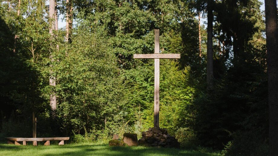 Holzkreuz, umgeben von Bäumen, mit einer steinernen Basis und einer Bank auf einer grünen Wiese im Wald.