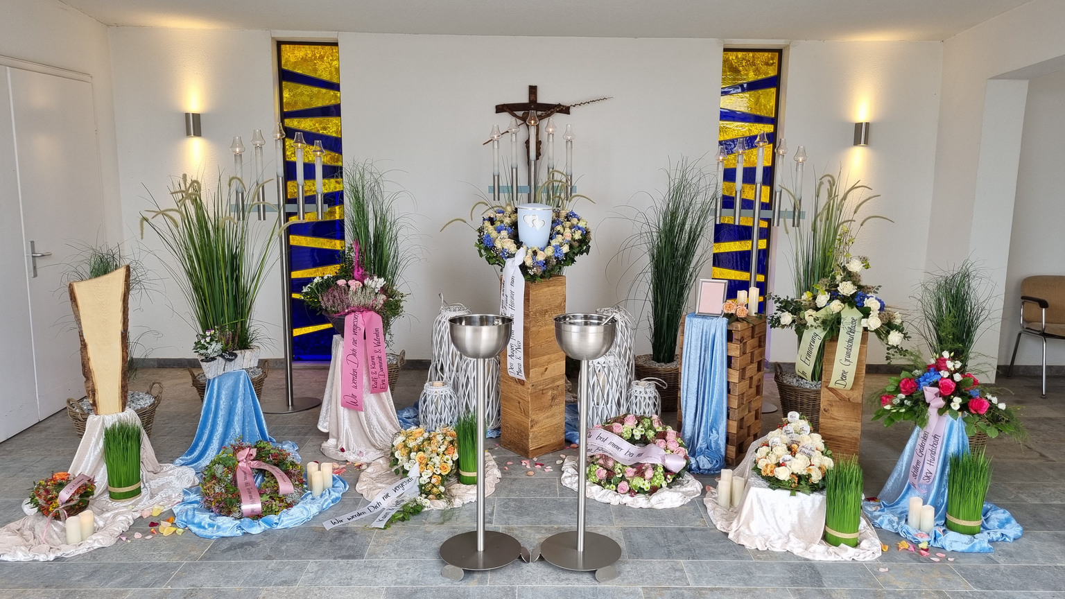 Dekorative Altarkerze und Blumenarrangements in einer Kirche, umgeben von farbigen Stoffen und einem bunten Fenster.