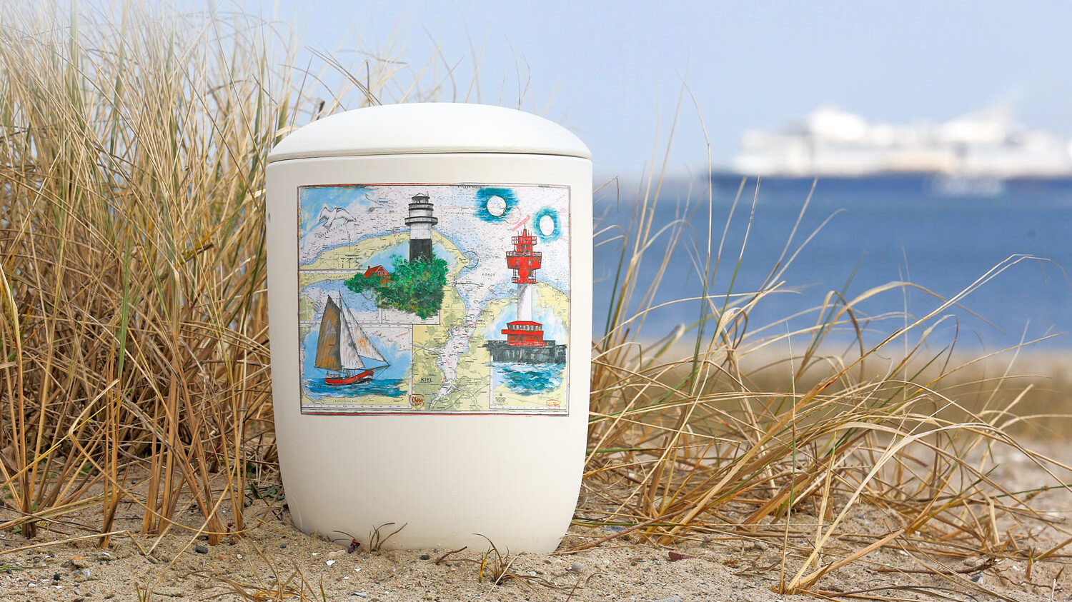 Abfallbehälter mit maritimer Illustration steht im Sand vor einer Küstenlandschaft und Meer im Hintergrund.