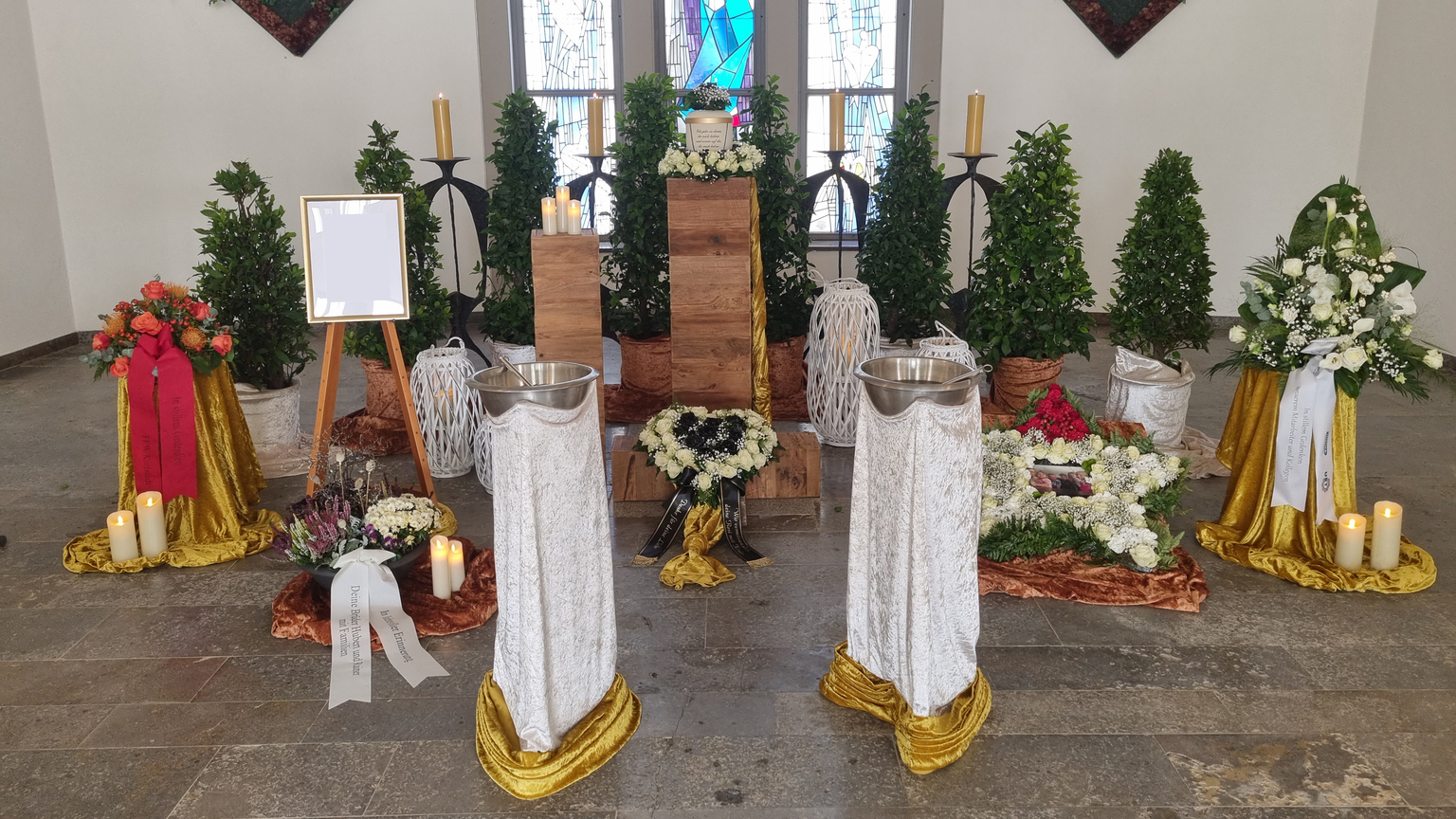 Dekorierte Trauerstätte mit Blumenarrangements, Kerzen, einem Kreuz und einem leeren Rahmen auf einem Holzpodest.