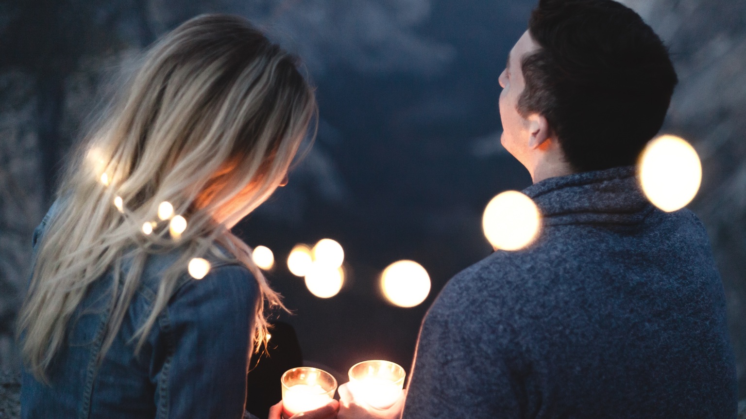 Paar hält leuchtende Kerzen, umgeben von funkelnden Lichtern, vor einer eindrucksvollen Berglandschaft.
