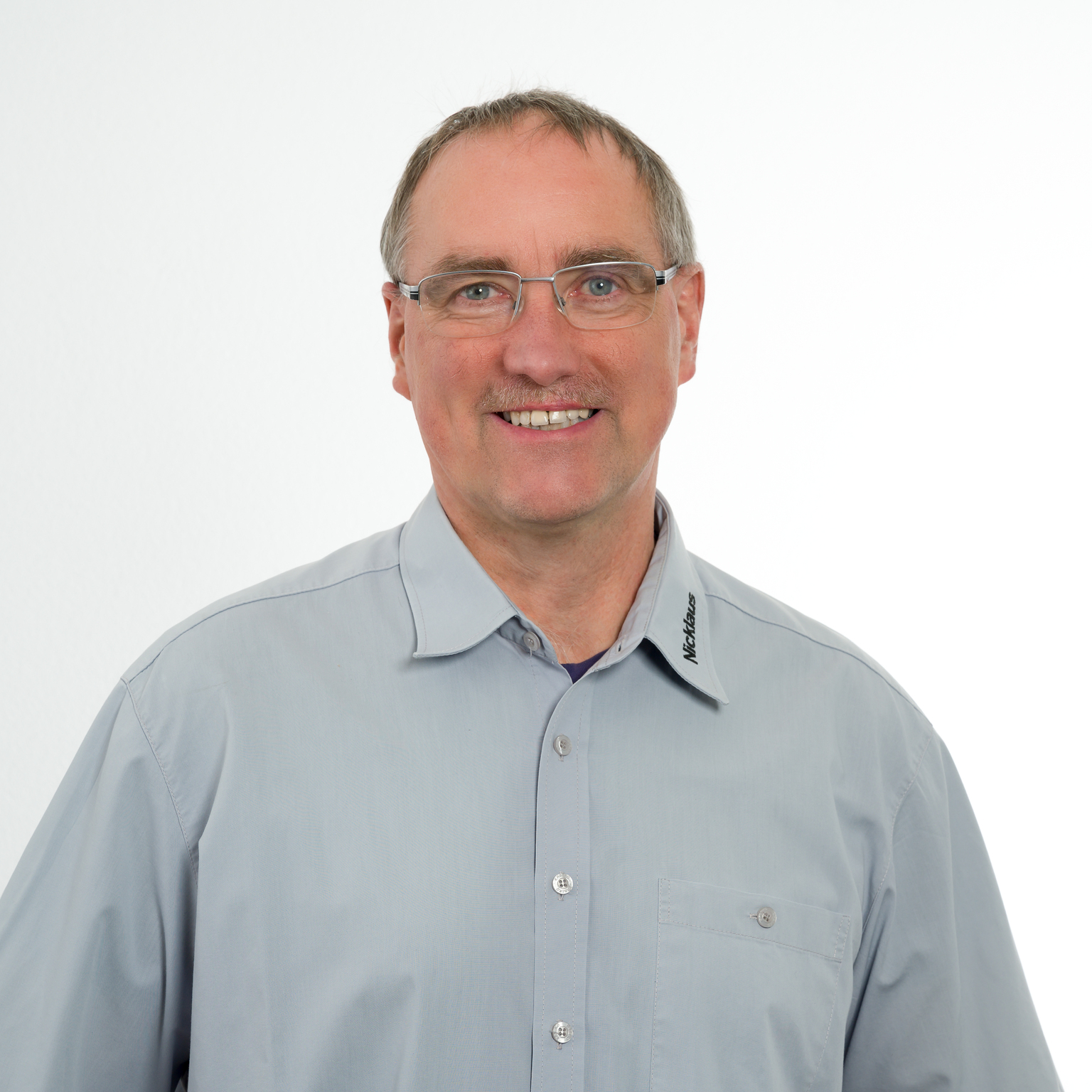 Porträt eines lächelnden Mannes mit Brille in einem grauen Hemd vor einem weißen Hintergrund.