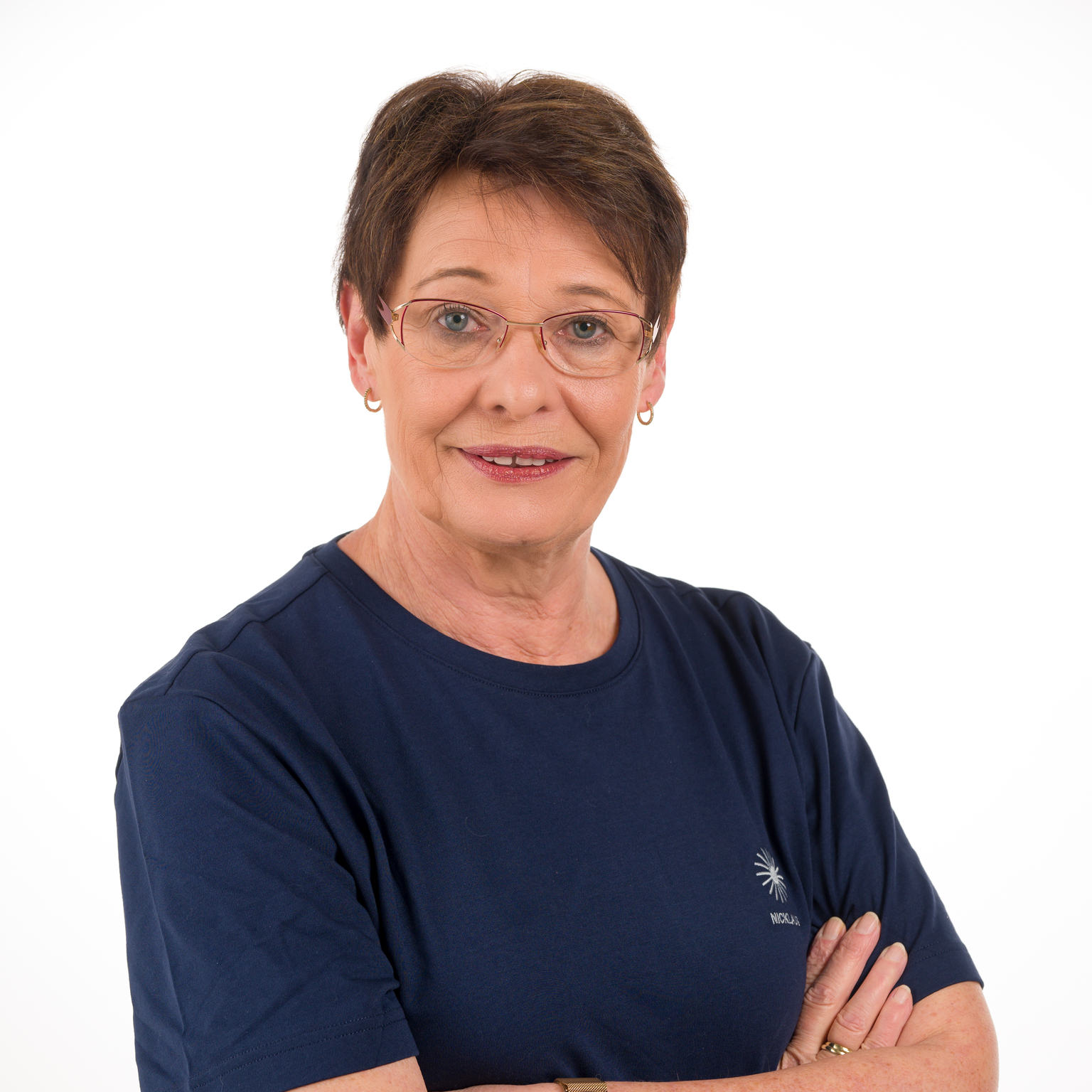 Porträt einer lächelnden Frau mit kurzen Haaren, Brille und blauer T-Shirt, die die Arme vor der Brust verschränkt.
