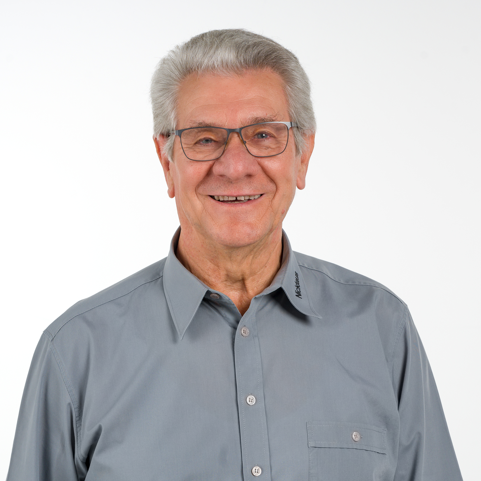 Älterer Mann mit Brille und grauem Hemd lächelt vor neutralem Hintergrund.