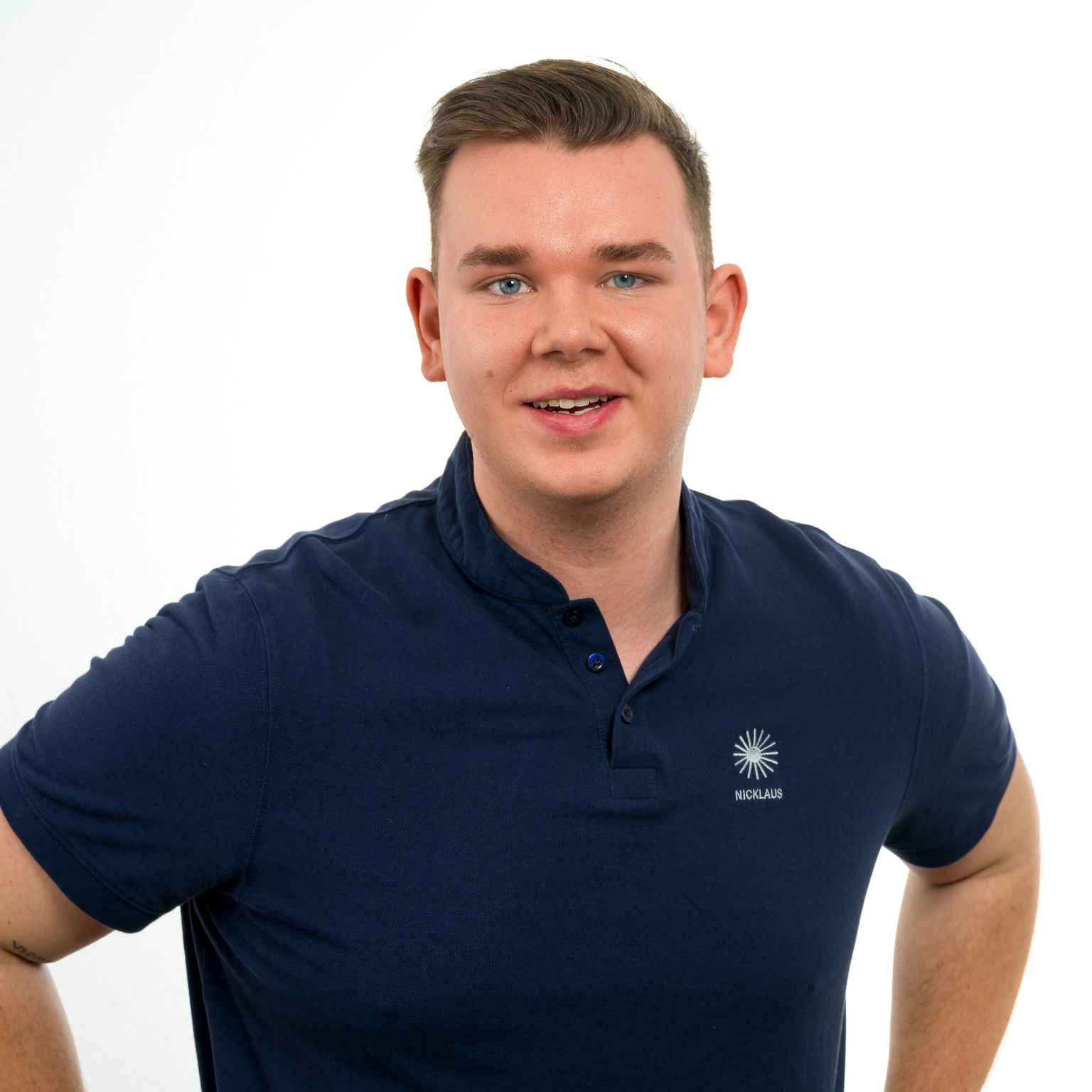 Junger Mann mit kurzem Haar und blauem Poloshirt lächelt freundlich vor einem weißen Hintergrund.