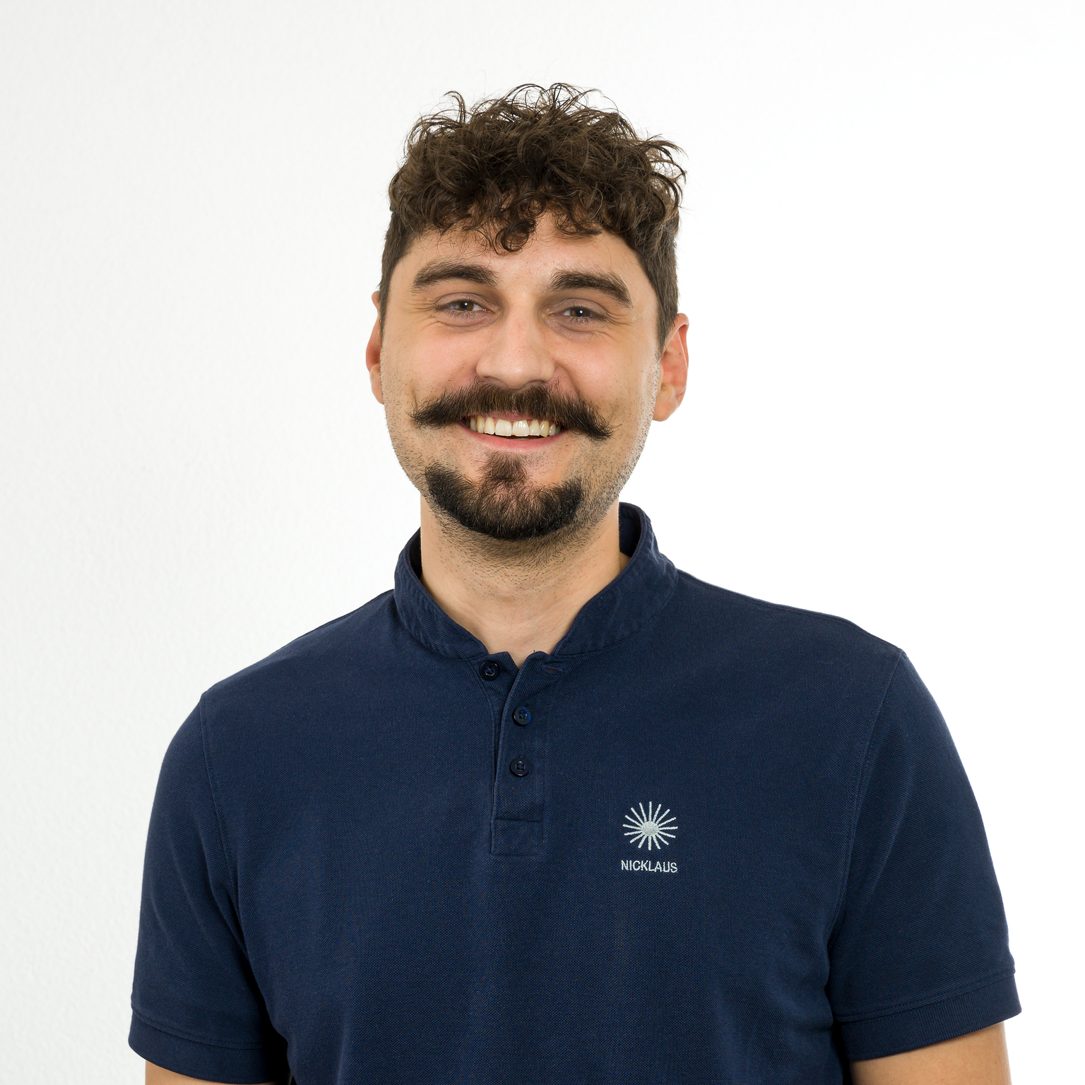 Porträt eines lächelnden Mannes mit lockigem Haar und Schnurrbart, der ein dunkelblaues Poloshirt trägt. Hintergründe sind weiß.