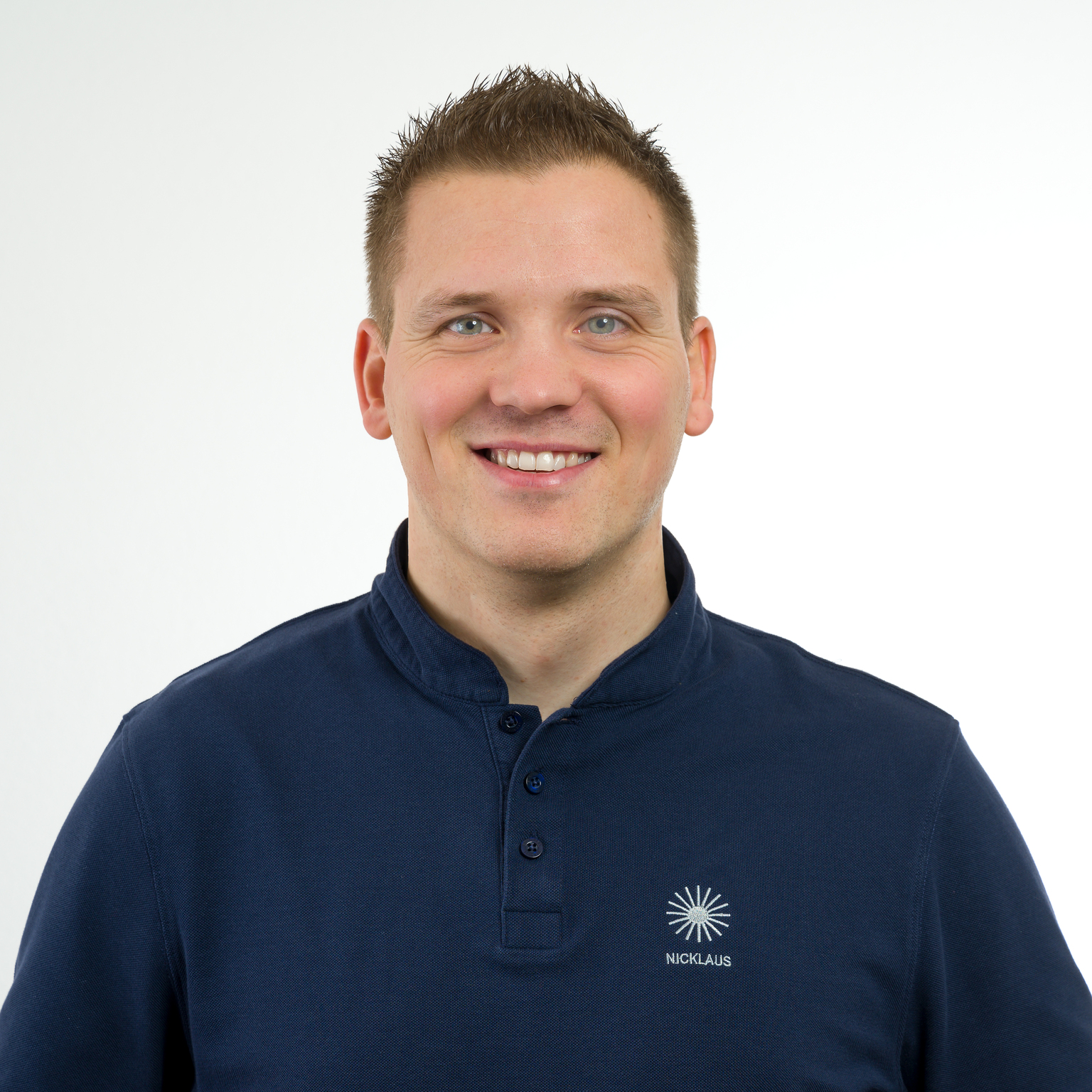 Porträt eines lächelnden Mannes mit kurzem, straffem Haar, der ein dunkles Poloshirt trägt, vor hellem Hintergrund.