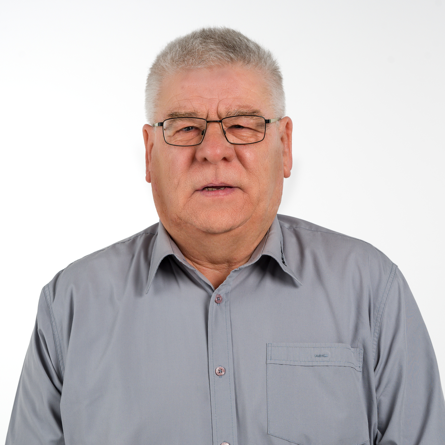 Porträt eines älteren Mannes mit Brille, in einem grauen Hemd vor einem weißen Hintergrund.