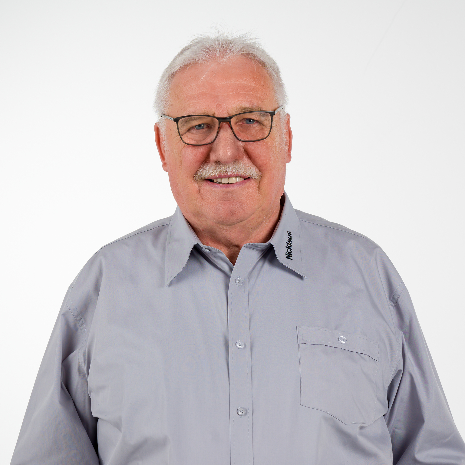 Portrait eines lächelnden Mannes mit Brille in einem grauen Hemd vor weißem Hintergrund.