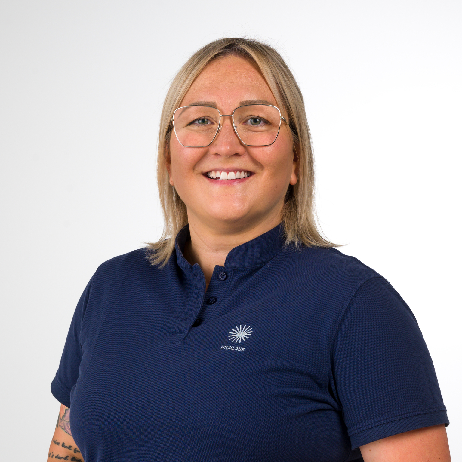 Portrait einer lächelnden Frau mit kurzen blonden Haaren, Brille und dunkelblauem Polohemd. Hintergrund ist hell.