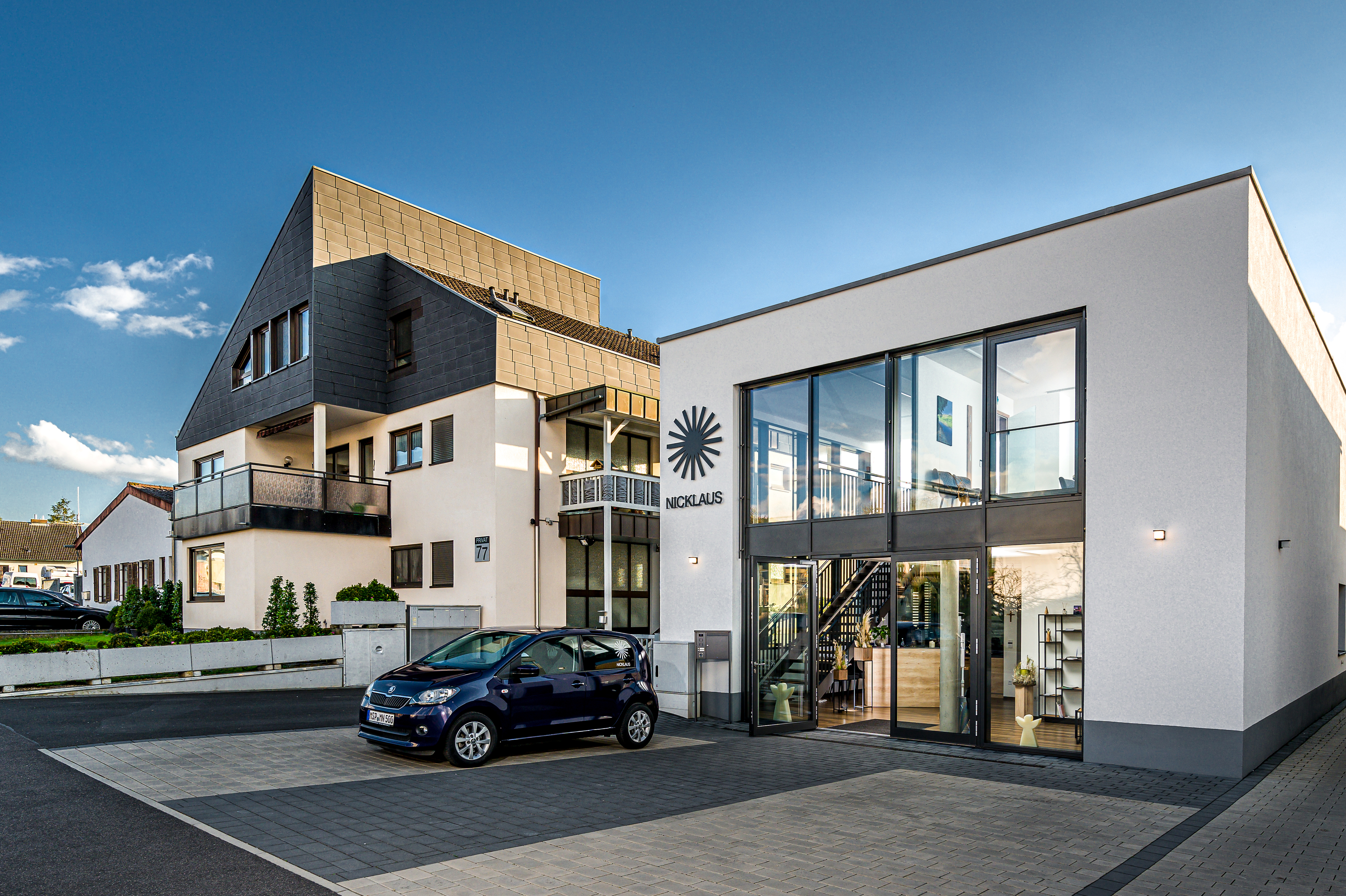 Moderne Gebäude mit Glasfassade und Treppe, davor ein blauer Smart und eine gepflegte Landschaft im Hintergrund.