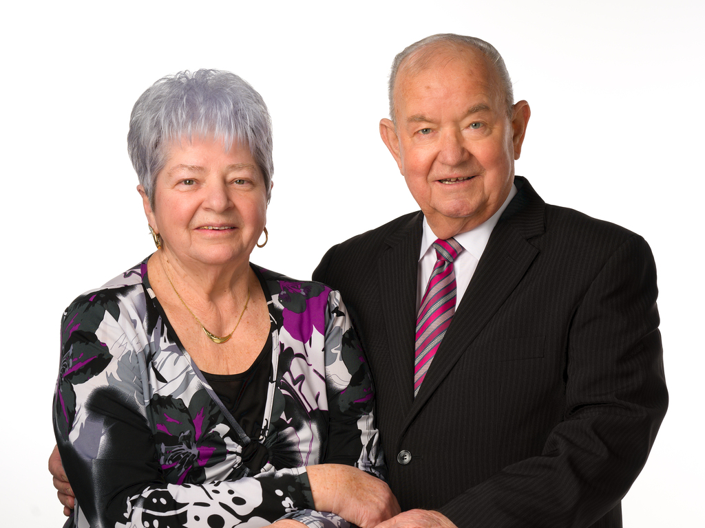 Ein älteres Paar posiert gemeinsam, lächelnd, vor einem hellen Hintergrund. Die Frau trägt ein blühendes Oberteil, der Mann ein Nadelstreifenanzug.