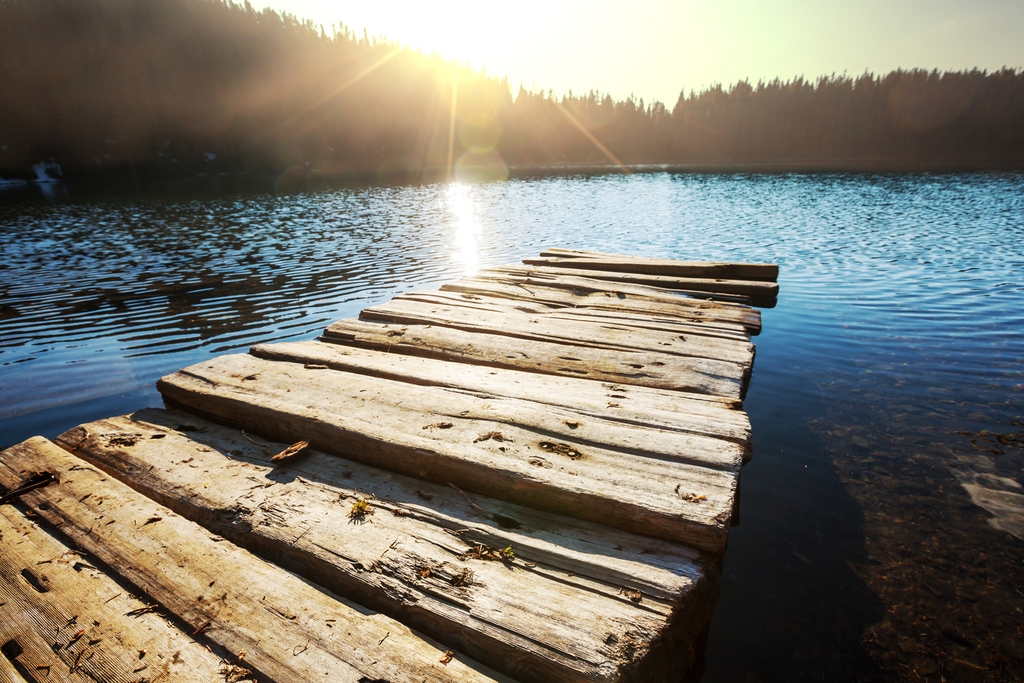 Holzpier am Ufer eines ruhigen Gewässers, beleuchtet von der Sonne, im Hintergrund eine bewaldete Landschaft.