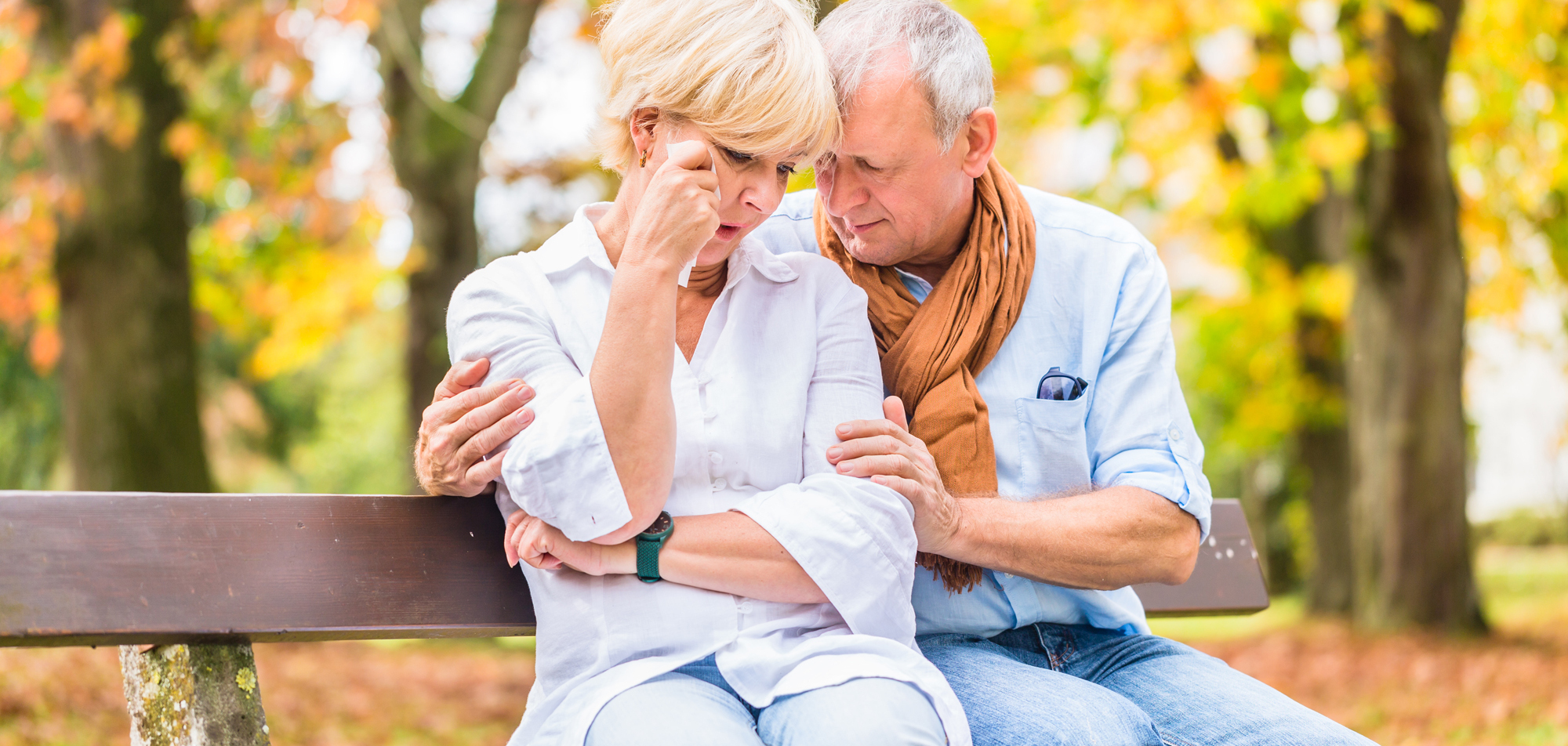 Ein älteres Paar sitzt auf einer Bank im Herbst, mit tröstender Körpersprache und ernsten Gesichtsausdrücken.